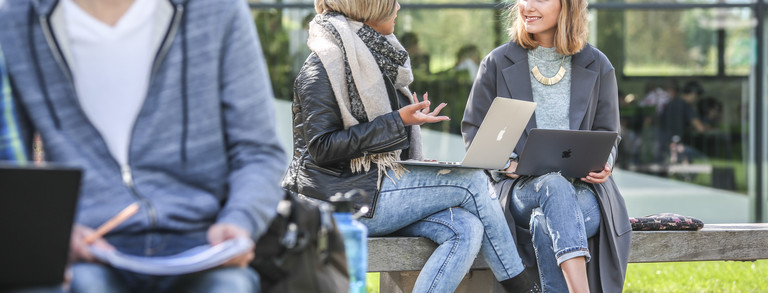 Zwei Studentinnen unterhalten sich draußen. Sie haben Laptops auf dem Schoß.